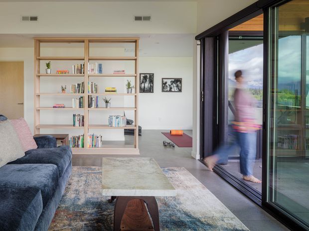 Bright contemporary living room with a deep blue sectional sofa, large patterned area rug, sculptural wooden coffee table, and open timber shelving filled with books and decor. Full-height dark-framed sliding glass doors open to an outdoor view, with a blurred person walking through the opening. The room has white walls, a light ceiling, polished concrete or grey flooring, and framed black-and-white artwork.