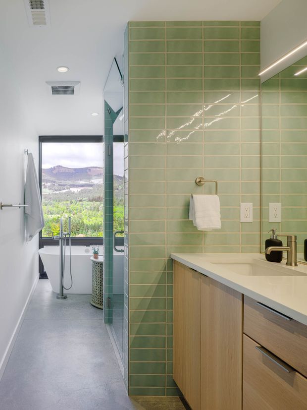 A bright contemporary bathroom with a double vanity in the foreground, pale timber cabinetry, a white benchtop, and a large wall finished in vertically stacked sage-green rectangular tiles with light grout. A glass shower partition leads toward a freestanding white bathtub beside a large window overlooking a green landscape. The room has a smooth light-grey floor, white ceiling with recessed lighting and ventilation grilles, brushed metal tapware, and warm integrated strip lighting reflected in a mirror.