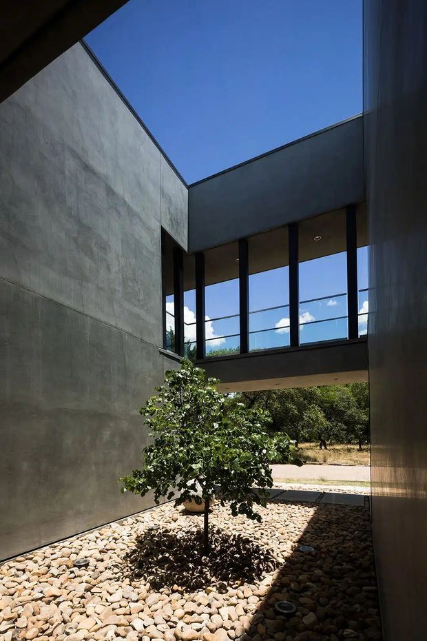 A narrow modern courtyard framed by tall smooth grey concrete or plaster walls, with a small tree planted in a bed of rounded stones. A dark minimalist upper-level bridge or wing spans the courtyard, featuring large floor-to-ceiling glazing and slender dark frames. Strong sunlight creates deep shadows, while blue sky and landscaped surroundings are visible beyond.