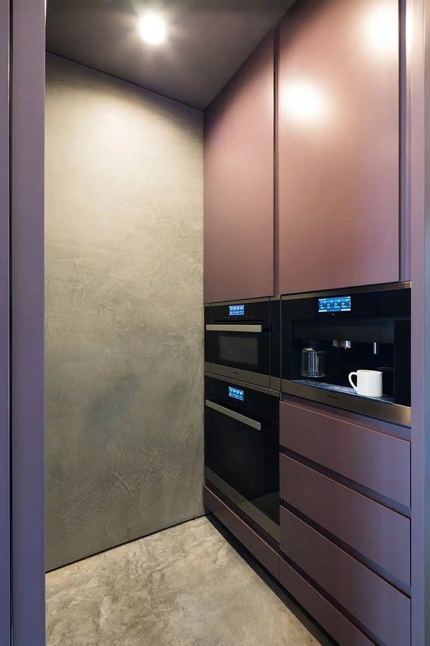 Compact, dark-toned built-in kitchen or beverage station with floor-to-ceiling mauve cabinetry, integrated black appliances, a built-in coffee machine, and a textured taupe-grey wall and floor. The space has a sleek, contemporary fitted design with concealed storage and warm recessed lighting.
