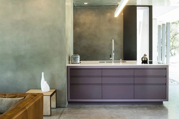 Contemporary kitchen interior featuring a large dark purple-grey island with integrated drawers and cabinetry, a stainless-steel sink and tap, and a dark stone or concrete benchtop. The room has textured grey concrete-look walls, a polished grey floor, black-framed glazing, and linear pendant lighting. A leather sofa and side table are partially visible in the foreground, suggesting an open-plan living area.