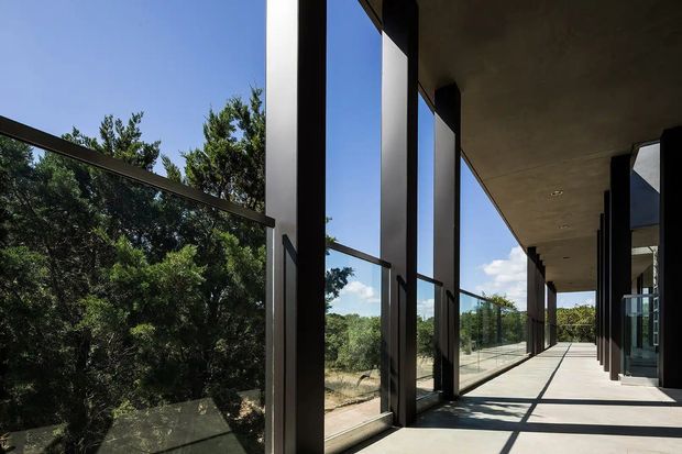 A long, covered exterior gallery or balcony with full-height dark-framed glazing and a minimalist concrete floor and ceiling. The transparent balustrade and windows open views toward dense green trees and a sunny landscape, while strong sunlight creates linear shadows across the walkway. The architecture is rectilinear, understated, and strongly connected to the surrounding outdoors.