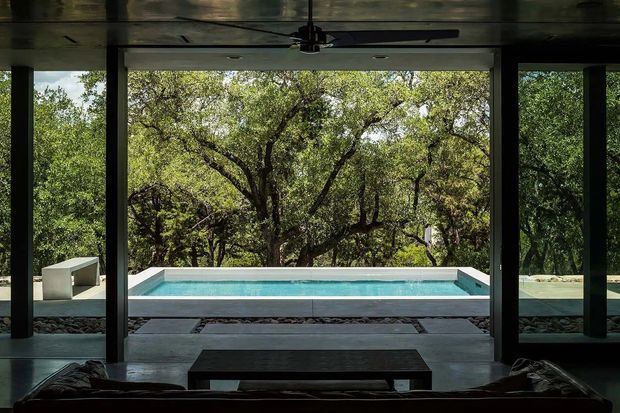 Dark, minimalist interior looking through extensive floor-to-ceiling glazing toward a rectangular swimming pool and dense wooded landscape. A wide overhanging ceiling with an integrated ceiling fan frames the view, while dark structural columns, polished concrete or stone paving, and low modern furniture create a strong indoor-outdoor connection.