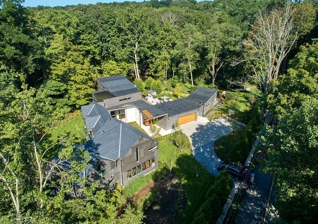 Aerial view of a multi-wing residential house nestled within dense woodland. The home has dark grey shingle-like cladding, multiple pitched and low-pitched roof forms, extensive glazing, and a central courtyard or driveway area with an attached garage featuring a warm timber door. The surrounding garden and mature forest dominate the setting.