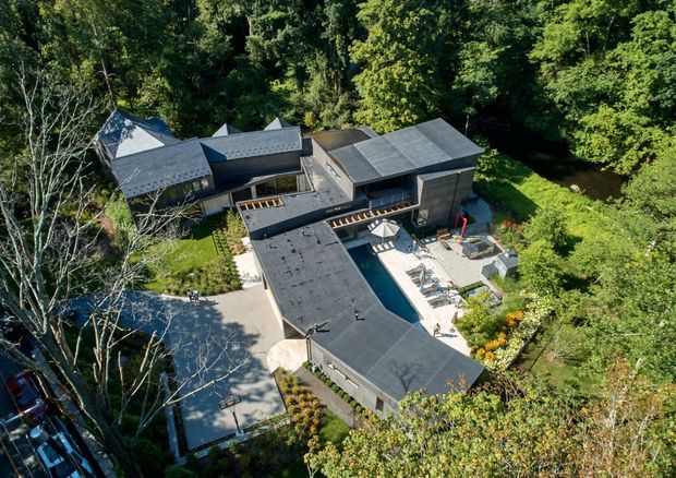 Aerial view of a large contemporary multi-wing residence nestled in dense woodland. The home has dark low-pitched roof planes, dark exterior walls, extensive glazing, a central courtyard with a long swimming pool, paved entertaining areas, landscaped garden beds, and a driveway or parking area along the left side.