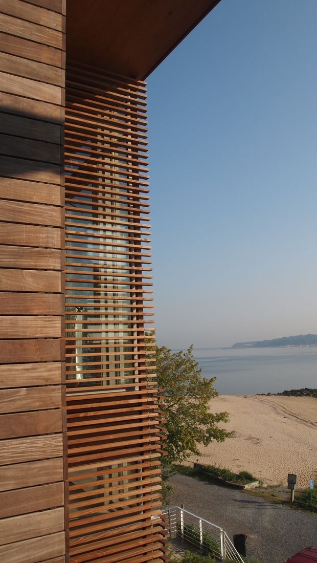 Vertical architectural exterior view of a contemporary waterfront building. The facade combines broad horizontal timber cladding with a prominent timber louvre or brise-soleil screen over tall glazed openings. A deep projecting roof canopy frames the upper portion, while a beach, calm water, and distant wooded shoreline are visible beyond the building. The composition emphasizes layered timber fins, glazing, and the building's relationship to the waterfront setting.