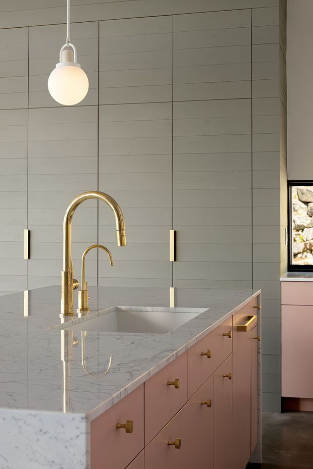 A refined contemporary kitchen interior dominated by a large marble-look island with an integrated undermount sink and sculptural polished brass gooseneck tap. The island features dusty pink cabinetry with brass pulls, while the backdrop is clad in pale grey horizontal and vertical-panel wall lining with slim brass wall lights. A white globe pendant hangs above, and a window at right reveals a stone exterior detail.