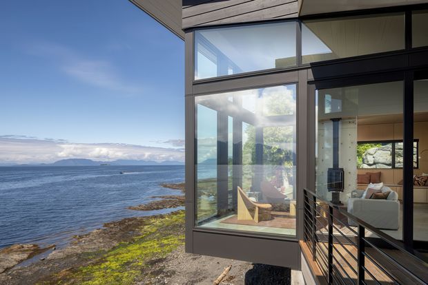 Exterior view of a contemporary coastal home projecting toward a rocky shoreline, with extensive floor-to-ceiling glazing, dark window frames, timber soffits, and a partially visible warm-toned living room inside. A railing-lined exterior walkway leads toward the glazed corner, and ocean and distant landforms form the background.