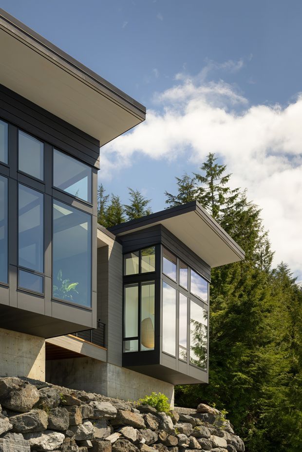 Exterior architectural photograph of a contemporary multi-level home set into a rocky, wooded site. The building features dark horizontal cladding, extensive black-framed floor-to-ceiling glazing, projecting box-like volumes, clean flat roof planes with pale soffits, and exposed concrete retaining walls above a natural stone embankment.