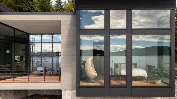 Exterior view of a contemporary multi-level home with dark framed floor-to-ceiling glazing, pale horizontal timber cladding, and a covered outdoor terrace. The glazing reveals a refined living area and bedroom, while reflecting dramatic clouds and surrounding mountain scenery. The composition emphasises strong geometric window grids, indoor-outdoor connection, and a cantilevered modern form.