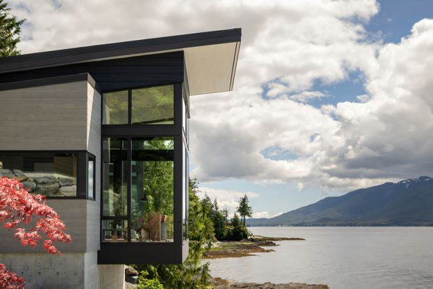 Exterior architectural photograph of a contemporary multi-level home projecting toward a calm waterfront. The building has dark horizontal roof edging, pale cladding, large black-framed floor-to-ceiling glazing, and a cantilevered glazed corner. A mountain-lined body of water and landscaped shoreline are visible beyond.