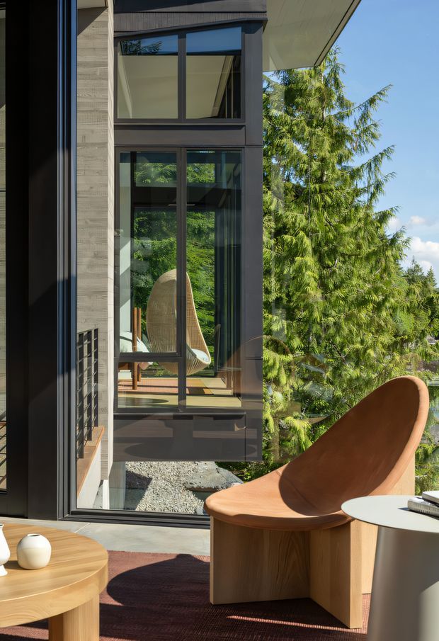 A sunlit contemporary interior beside a tall glazed facade, featuring floor-to-ceiling dark-framed windows, warm timber furniture, a sculptural tan lounge chair, and views of mature greenery outside. The image emphasizes the connection between the refined living space and the surrounding landscape.