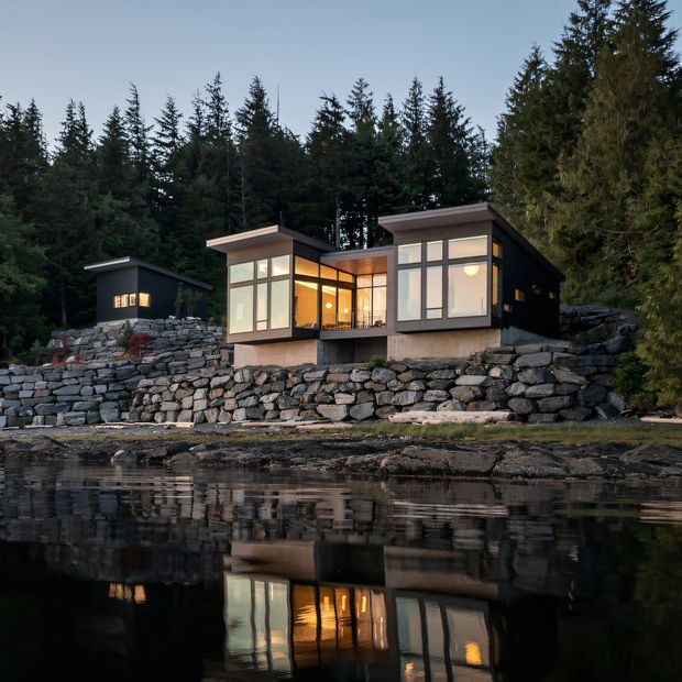 A modern flat-roofed house sits on a rugged stacked-stone retaining wall beside calm reflective water, surrounded by dense evergreen forest. The dark-clad structure has expansive floor-to-ceiling glazing, warm interior illumination, and a smaller matching outbuilding to the left. The image is a dusk exterior architectural photograph with a strong waterfront setting and reflected house lights.