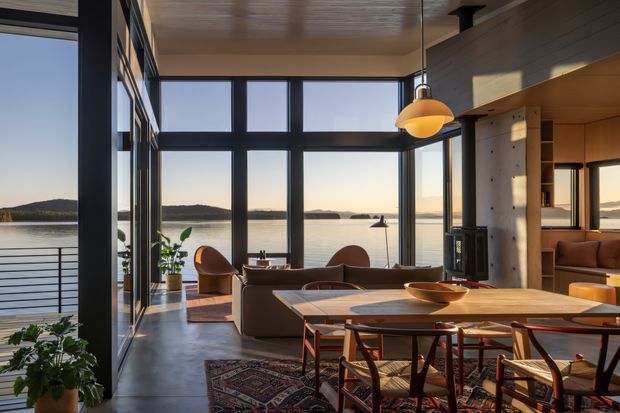 A warm, contemporary open-plan dining and living space overlooking calm water through expansive floor-to-ceiling glazing. The room features a large timber dining table with sculptural wooden chairs, a low upholstered sofa, rounded lounge chairs, a patterned rug, a suspended pendant light, timber-lined ceiling and walls, and dark-framed windows. A balcony with metal balustrading and landscaped plants is visible outside.