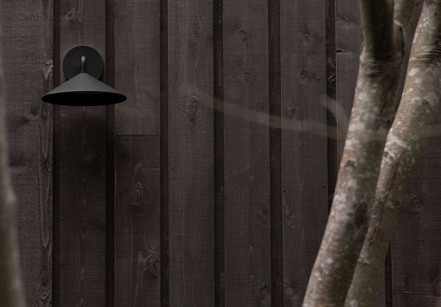 Close-up of a dark timber-clad wall made from vertical weathered boards, featuring a minimalist black wall-mounted dome light. Tree trunks partially obscure the right side, creating a moody, textural architectural detail.