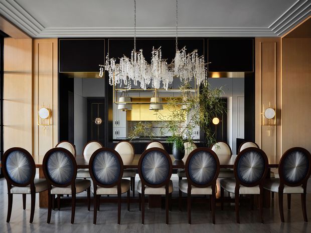 A sophisticated formal dining room with a long dark wood dining table surrounded by upholstered round-back dining chairs. The room features dramatic black wall panels, warm timber wall cladding, layered decorative ceiling moulding, brass wall sconces, and a large cascading crystal chandelier. A mirrored or reflective rear wall and built-in display or bar joinery add depth, while greenery provides a natural accent.