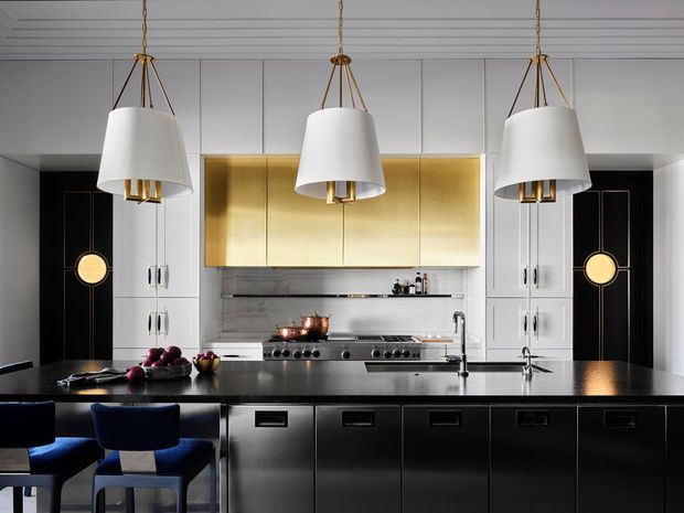 A sophisticated kitchen with a large dark island and integrated black cabinetry, white paneled walls, a stainless-steel cooking range, and a striking brushed-gold overhead cabinet feature. Three oversized white pendant lights hang above the island, while black-and-gold decorative wall panels and blue upholstered stools add contrast. The space has a refined, high-contrast contemporary Art Deco-inspired aesthetic.