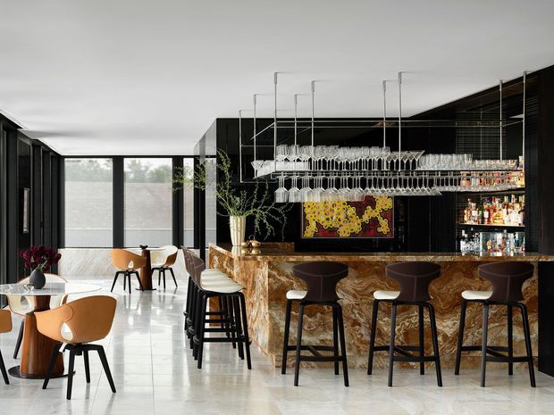 A sophisticated contemporary bar and dining space with a long sculptural marble bar, dark ribbed cabinetry, extensive bottle storage, and rows of suspended wine glasses. Warm timber and tan leather dining chairs contrast with the black joinery, while full-height glazing brings in soft natural light. The polished light stone floor and dramatic patterned bar front create a refined hospitality-style interior.