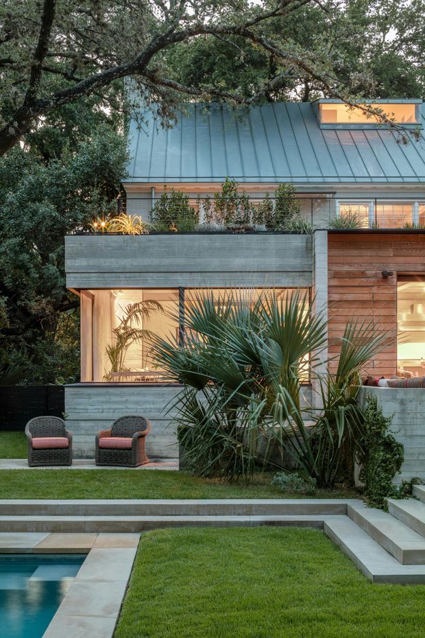 A landscaped residential backyard at dusk, featuring a modern house with a steep standing-seam metal roof, timber and grey concrete or cement-finished walls, expansive warm-lit glazing, and a raised horizontal window. A rectangular swimming pool, stepped concrete paving, manicured lawn, planted garden beds, and two outdoor lounge chairs occupy the foreground. Mature trees frame the house, while spiky architectural planting provides a strong landscape feature.