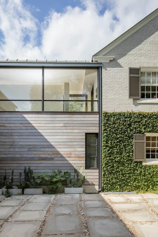 Exterior architectural photograph showing a contemporary glazed extension adjoining a light-painted brick house. The extension has a low metal roof, large black-framed glazing, horizontal timber cladding, and a narrow vertical window. The original house features a steep gabled roof, white brickwork, timber-framed windows with dark shutters, and a wall covered in climbing greenery. A paved courtyard with large concrete slabs and gravel joints occupies the foreground, with potted plants along the extension.