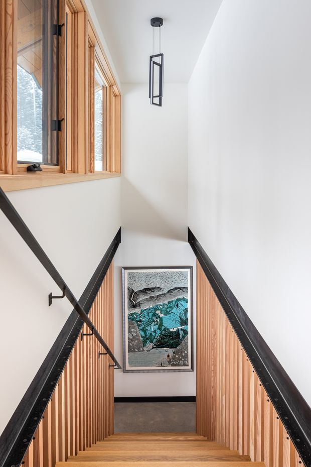 A bright, narrow stairway descending through a contemporary interior. The staircase has warm timber treads and vertical timber slat balustrade panels, white walls, dark metal handrails and trim, and a framed artwork at the lower landing. Timber-framed windows bring in natural light, while a simple suspended geometric pendant adds a modern detail.