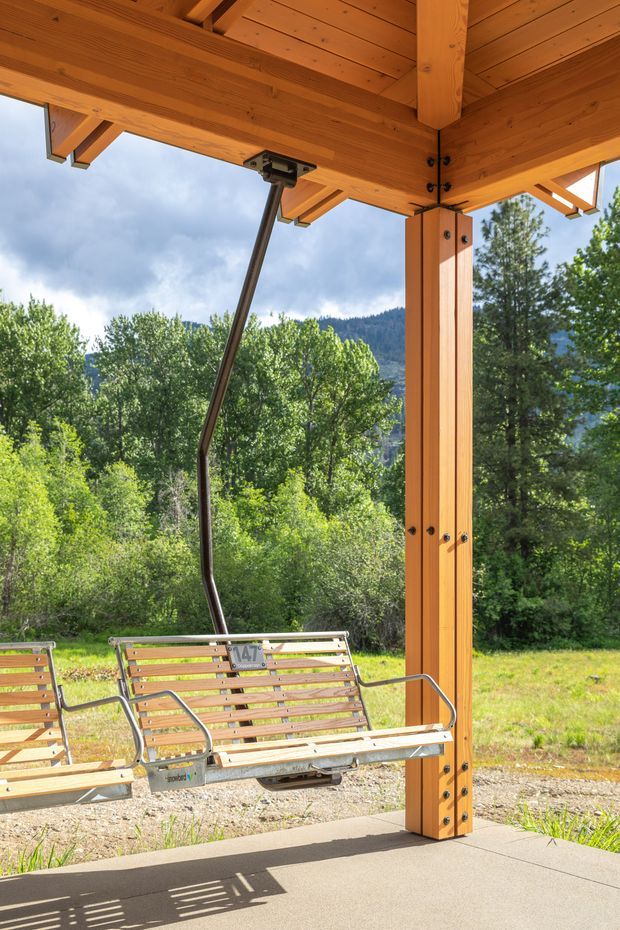 Open-air chairlift loading station beneath a large exposed timber canopy, with heavy timber posts, beams, rafters and visible metal structural connections. Wooden ski-lift seats are suspended in the foreground, overlooking a wooded mountain landscape. The image is bright and outdoors, focused on the covered lift structure and its robust timber construction.