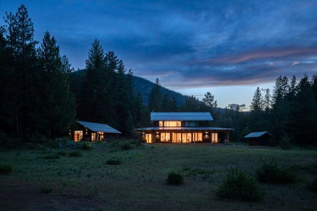 A contemporary single-storey house complex glowing with warm interior light at dusk, set in a forested mountain landscape. The main building has extensive glazing, a low-pitched metal roof with a raised clerestory-like central volume, and dark timber or metal exterior elements. Smaller detached structures sit to either side across an open meadow.