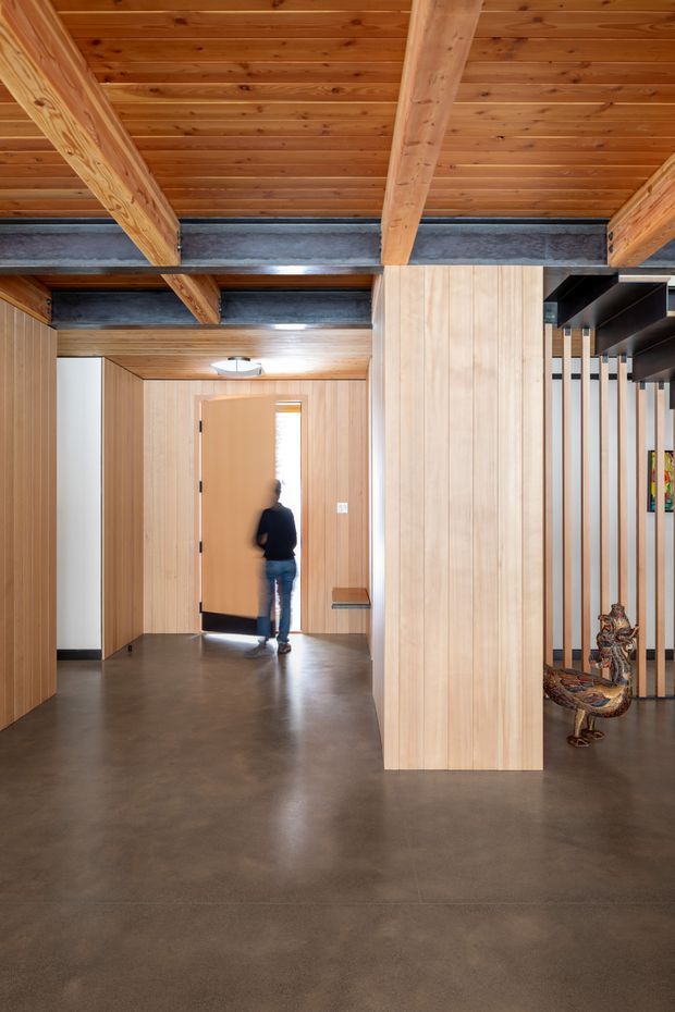 A warm, contemporary timber-lined interior hallway or entry space with exposed timber ceiling boards and substantial timber beams supported by dark steel framing. Full-height pale timber wall panelling and a central timber-clad partition create a series of circulation zones. The polished dark concrete floor contrasts with the extensive wood surfaces, while vertical timber slats and dark open shelving screen a display area on the right. A person is visible near an open doorway in the background.