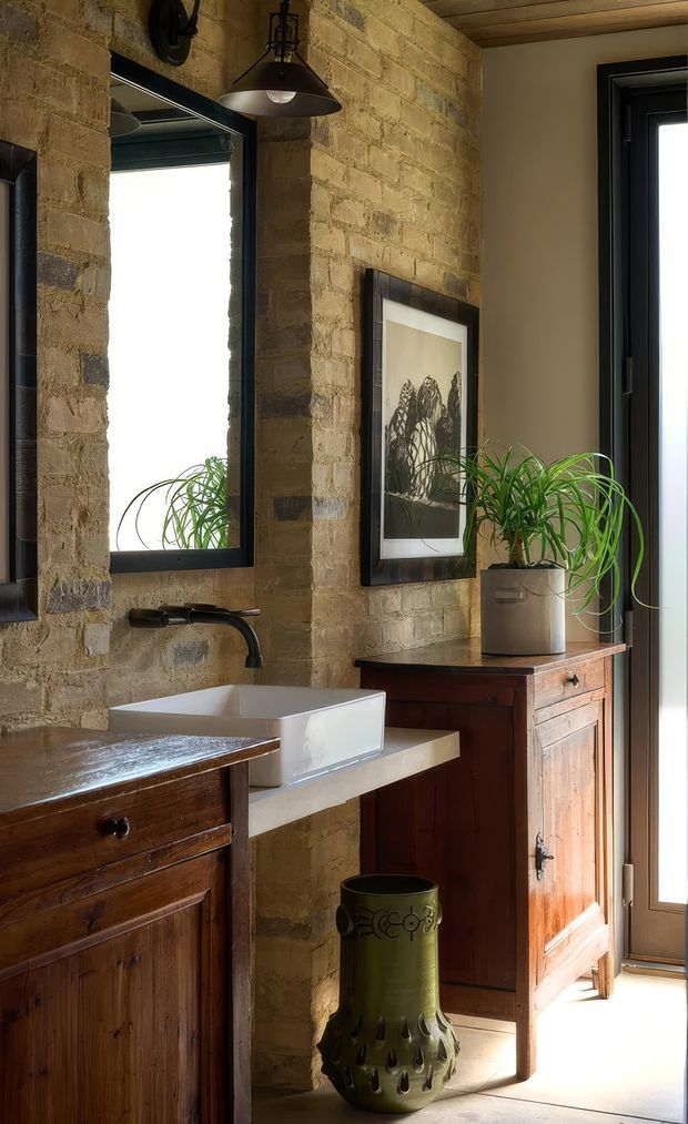 Warmly lit rustic bathroom featuring exposed tan masonry block walls, dark-framed mirrors and glazing, a white vessel sink on a pale stone or solid-surface counter, dark timber vanity cabinetry, and a freestanding wooden cabinet with a potted plant. Black industrial-style wall and pendant lighting, framed artwork, and natural timber ceiling add character. The room has a warm, earthy, vintage-industrial atmosphere.
