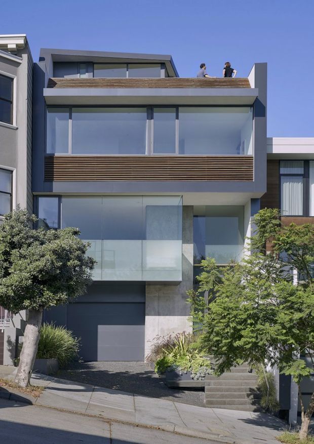 Street-facing view of a narrow multi-storey contemporary townhouse with a strong rectilinear form. The facade combines large floor-to-ceiling glazing, grey rendered or concrete-like surfaces, dark metal-framed windows, horizontal timber privacy screens, projecting balconies, and a recessed garage and entry. Mature trees and planting partially screen the lower levels, while two people are visible on the rooftop terrace. The building sits on a sloping urban street.