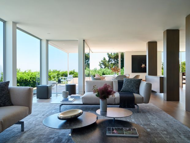 Bright, spacious contemporary living room with floor-to-ceiling glazing opening toward a lush outdoor setting. The room features a large pale sectional sofa, sculptural lounge chairs, layered round coffee tables, a broad grey area rug, dark timber or walnut-toned vertical columns, and a wall-mounted television. The open-plan area extends toward a dining or sitting zone in the background, with strong indoor-outdoor visual connection and abundant natural light.