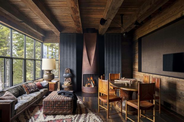 Rustic, dark-toned living and dining room with extensive exposed timber ceiling beams and timber wall cladding. Large black-framed windows overlook trees, while a leather sofa, patterned ottoman, dining table and chairs, built-in television, and sculptural central fireplace create a warm lodge-like interior.