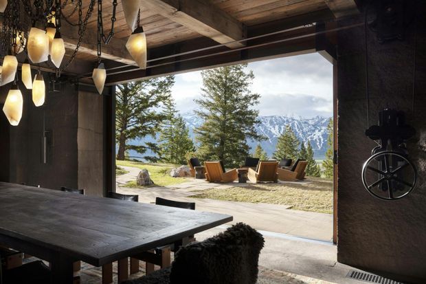 Rustic industrial-style dining or entertaining space opening through a wide glazed garage-like opening to an outdoor seating area and dramatic mountain landscape. The interior features a large dark timber dining table, exposed timber ceiling beams, dark textured walls, and sculptural pendant lighting suspended from chains. A prominent wheel-like wall feature adds a workshop-inspired detail.