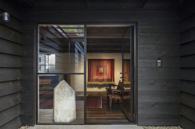 Exterior view of a dark, horizontally clad building facade with a large black-framed glazed entry or window. Through the glazing, a warm timber-lined interior with exposed beams, hardwood flooring, a rug, seating, and a prominent illuminated crystal-like decorative object is visible. The composition emphasizes the dark exterior cladding and contrast between the cool facade and warm interior.