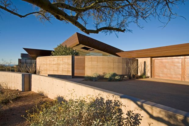 Exterior view of a low-slung contemporary residence with dramatic angular roof planes, warm timber cladding and garage doors, dark-framed glazing, and exposed concrete retaining walls. The house is set behind a landscaped forecourt with native-style planting and a broad paved driveway.