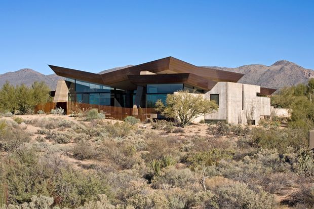 Exterior view of a low, contemporary desert residence set within sparse scrubland and rugged mountains. The home features angular projecting roof planes, extensive dark-framed glazing, warm timber soffits and cladding, and pale textured masonry or concrete walls. Native desert planting and rocky terrain surround the building.