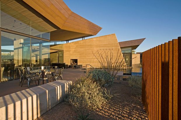 Exterior view of a contemporary, low-profile building with expansive floor-to-ceiling glazing, warm timber-clad angular roof and wall forms, and a paved outdoor terrace furnished with tables and chairs. A rust-toned vertical timber privacy screen and drought-tolerant landscaped planting frame the courtyard-like setting.