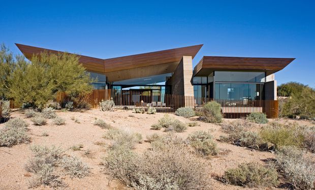 Single-storey contemporary house set in a sparse desert landscape, with broad angular projecting roof planes, extensive floor-to-ceiling glazing, warm timber-clad soffits and facade elements, and a dark structural frame. Native drought-tolerant shrubs and gravelly sandy landscaping surround a timber-railed outdoor terrace.