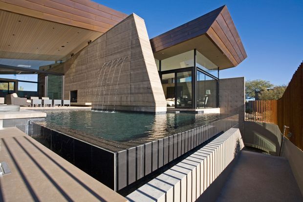 A striking modern residential exterior centered on a raised reflecting or swimming pool beside a sculptural concrete wall with water feature. The house combines extensive black-framed glazing, sharp geometric volumes, warm copper-toned metal roof cladding, and timber-lined soffits. A paved terrace and stepped pool edge occupy the foreground, with a timber privacy fence and distant landscape beyond.