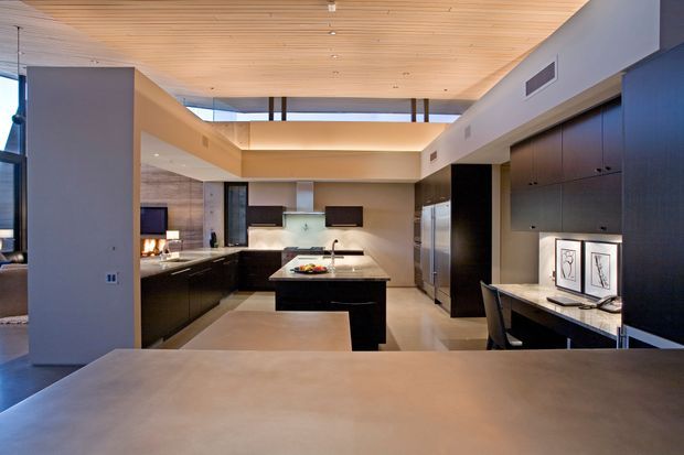 A spacious contemporary open-plan kitchen viewed across a broad stone island or benchtop. The room features dark built-in cabinetry, stainless-steel appliances, pale stone surfaces, a timber-lined ceiling, and a raised clerestory-style glazed opening that brings in natural light. A small integrated desk or study nook with framed artwork is visible at right.
