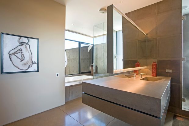 Contemporary bathroom with a floating double vanity in a warm grey finish, integrated basins, a large backlit mirror, glass-enclosed bathtub area, and floor-to-ceiling glazing. The space has clean geometric lines, polished concrete-like surfaces, and restrained decorative accents.