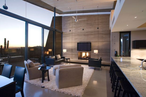 Open-plan contemporary living and kitchen area with a dramatic double-height volume, floor-to-ceiling glazing, and a timber-lined ceiling. The living room centres on a wall-mounted television and fireplace against a textured concrete feature wall, with lounge seating arranged on a pale rug. A large stone kitchen island with dark bar stools occupies the right side, while pendant lights and dark-framed glazing reinforce the modern architectural character.