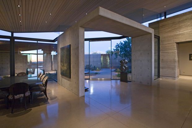A sophisticated open-plan interior with exposed board-formed concrete walls and portal-like structural elements, polished light flooring, a slatted timber ceiling, and extensive black-framed floor-to-ceiling glazing. A dining area occupies the left side, while the central space opens toward a landscaped courtyard and pool-like reflective water feature. The restrained palette and dramatic evening lighting create a refined, gallery-like atmosphere.