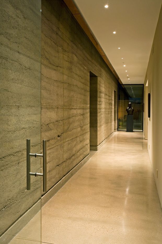 A long, narrow minimalist interior hallway with a polished concrete floor and exposed textured concrete block walls. A frameless glass door with stainless steel pull handles occupies the foreground, while recessed ceiling downlights lead toward a glazed end wall and a dark pedestal displaying a sculptural bust. Warm concealed timber detailing and neutral lighting create a refined, gallery-like atmosphere.