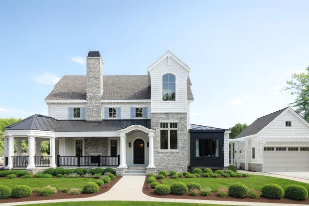 Front exterior view of a large, traditionally styled multi-storey house with a prominent gabled central bay, light stone chimney and lower facade, white weatherboard cladding, dark standing-seam metal porch roof, covered veranda with columns and railings, and an attached garage wing. Formal hedged landscaping and a central concrete path lead to the entry.