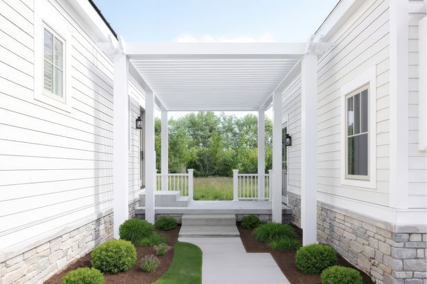 Bright exterior view of a covered entry walkway between two white-clad building wings. The passage has horizontal weatherboard siding, stone veneer bases, white-framed windows, a white pergola-style roof, and a concrete path bordered by low shrubs and ornamental grasses.