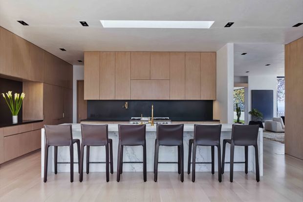 A refined contemporary kitchen viewed from the dining side, featuring a large marble island with seating for six, full-height light timber cabinetry, a dark backsplash, integrated appliances, and warm timber wall panelling. Recessed ceiling lights and a long skylight provide even illumination, while an adjacent living area is visible beyond the kitchen.