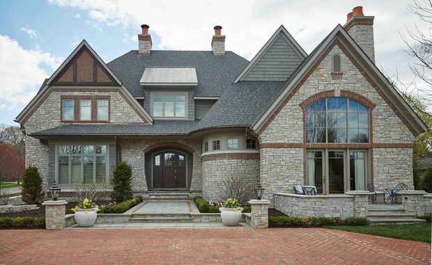 Front exterior view of a large, multi-gabled stone-clad home with steep pitched roofs, prominent arched window, dormer, multiple chimneys, timber and brick detailing, and a formal brick-paved driveway. The residence has a substantial traditional character with landscaped entry planting and a raised terrace.