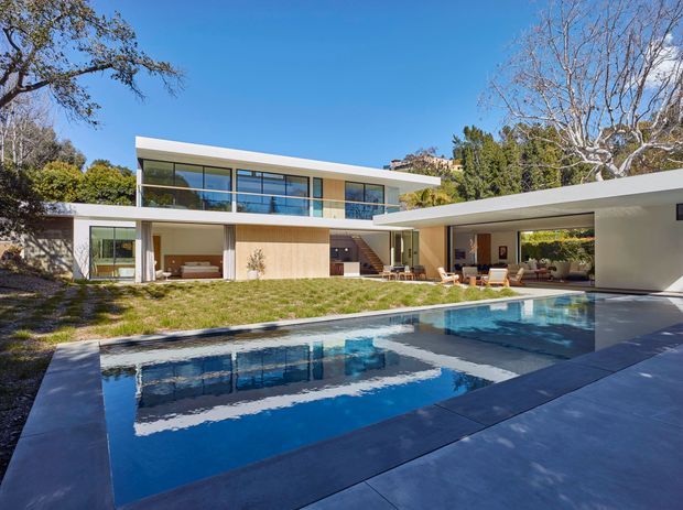 Exterior view of a large modern multi-wing residence arranged around a swimming pool. The home has crisp white rendered walls, extensive floor-to-ceiling glazing, a low-pitched flat roof, warm timber wall cladding, and a covered outdoor entertaining area with lounge and dining furniture. The pool and broad paved terrace dominate the foreground, with mature trees surrounding the property.