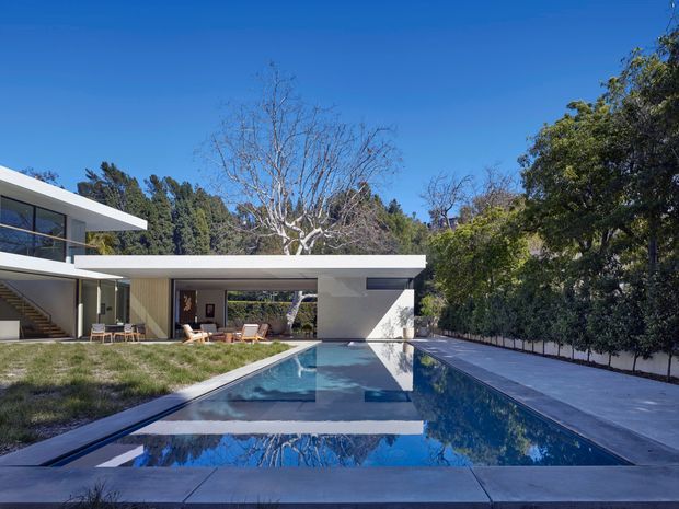A contemporary, low-profile residence set within a landscaped suburban garden, viewed from beside a long rectangular swimming pool. The home has crisp white flat-roofed volumes, extensive glazing, a covered outdoor living area, and an upper-level terrace. Pale paving surrounds the pool, while mature trees and hedges provide privacy. The scene is bright and strongly geometric, with blue sky and water reflections.